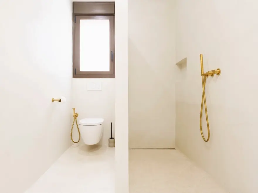 Aseo con microcemento blanco, WC suspendido y ducha con grifería dorada en vivienda de Elche