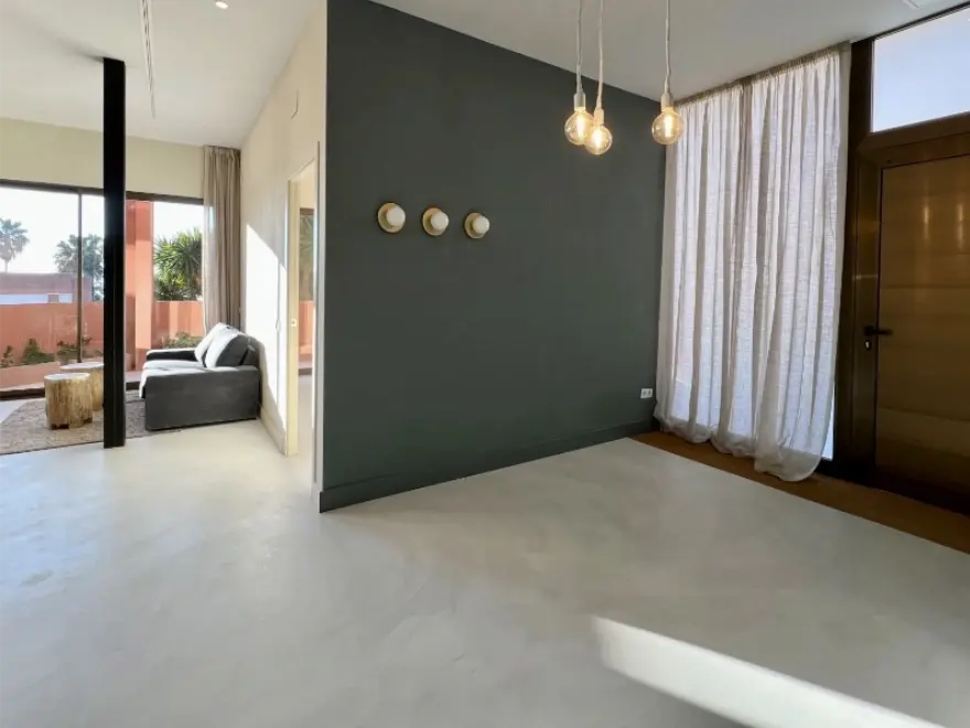 Salón con pared de microcemento verde oscuro, suelo blanco y cortinas blancas en vivienda de Elche