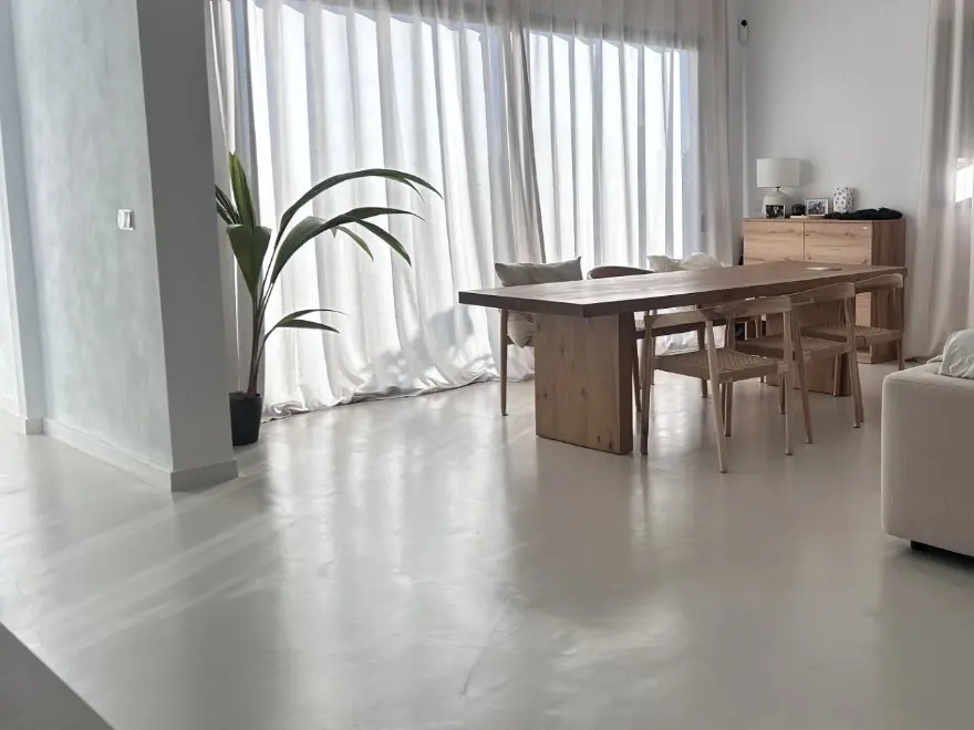 Comedor con suelo de microcemento gris claro, mesa de madera y cortinas blancas en vivienda de Elche