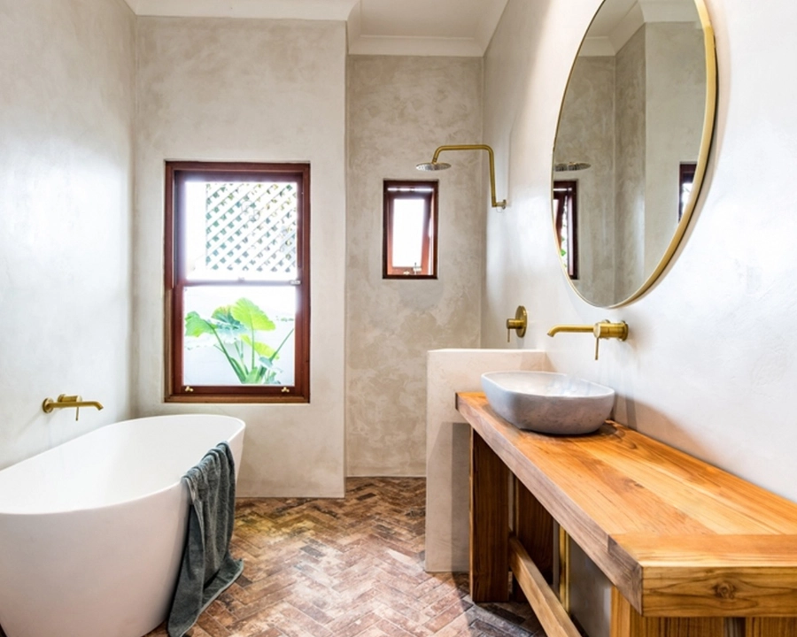 Baño con paredes de microcemento gris claro, bañera exenta, encimera de madera y griferías doradas en Elche