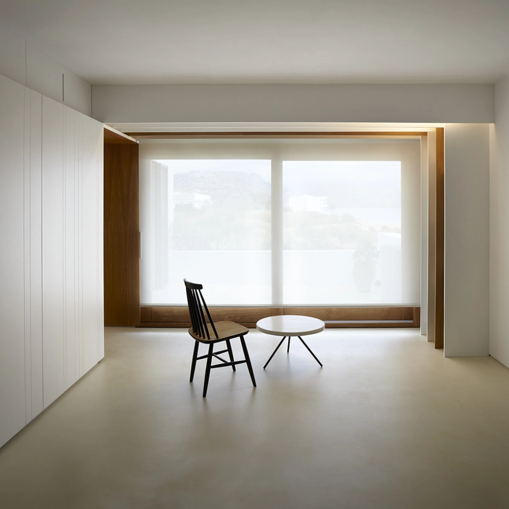 Salón minimalista con suelo de microcemento beige claro, armarios blancos y ventanal con luz natural en Elche