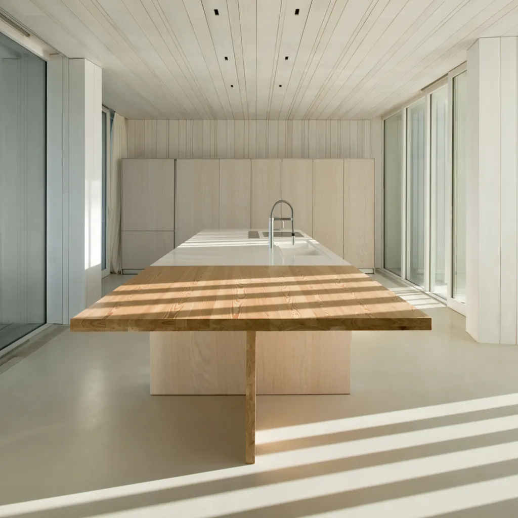 Cocina minimalista con suelo de microcemento gris claro, isla blanca y mesa de madera maciza con luz natural en Elche