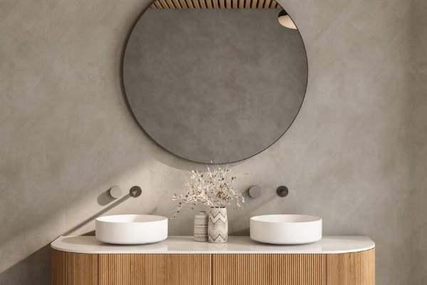 Detalle de baño con pared de microcemento gris topo, doble lavabo sobre mueble de madera y espejo redondo en Elche