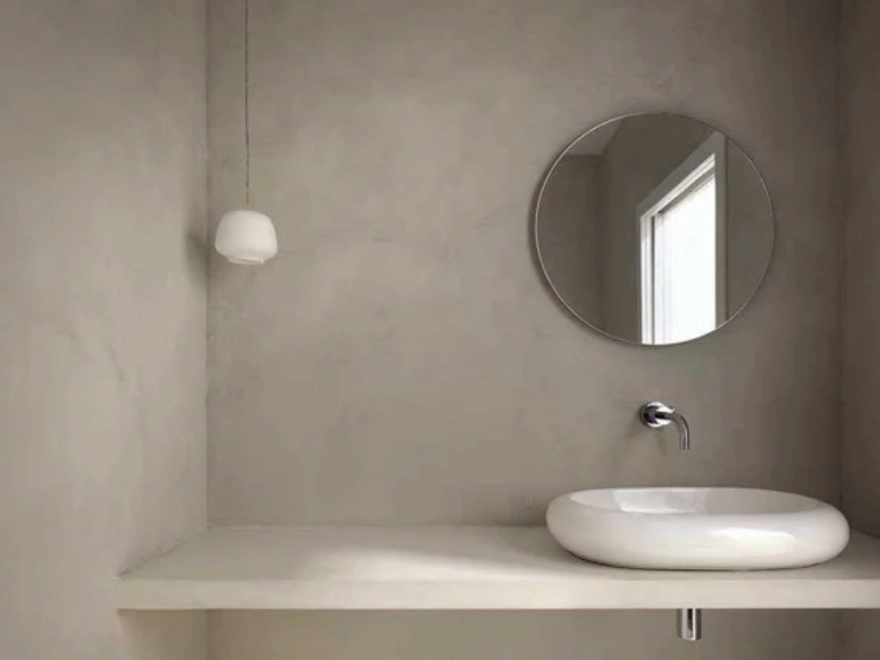 Baño minimalista con microcemento gris topo en paredes y encimera, lavabo sobre encimera y espejo redondo en Elche